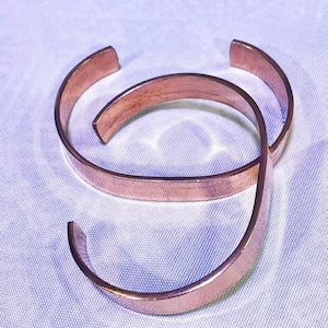 May include: Three copper-coloured open cuff bracelets. The bracelets are made of a flat, polished metal and have a simple, minimalist design. The bracelets are arranged on a white surface.