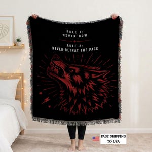 Könnte beinhalten: Eine schwarze Webdecke mit einer roten Grafik eines knurrenden Wolfskopfes. Der Text lautet: RULE 1: NEVER BOW, RULE 2: NEVER BETRAY THE PACK. Die Decke hat einen gefransten Rand und ein kontrastreiches Design mit strahlenförmigen Linien.