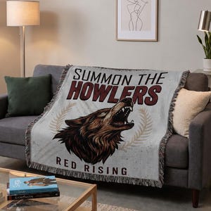 Könnte beinhalten: Eine gewebte Wohndecke mit einer Grafik eines heulenden Wolfskopfes in Braun- und Schwarztönen. Der Text lautet "SUMMON THE HOWLERS" in großen dunkelroten Buchstaben über dem Wolf und "RED RISING" in kleineren schwarzen Buchstaben unter dem Motiv.