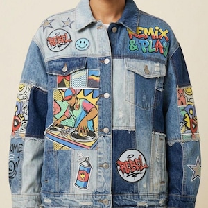 May include: A patchwork denim jacket featuring various graphic prints and patches. The design includes a DJ illustration, spray paint can, stars, smiley faces, and the text "REMIX & PLAY" and "REBEL". The jacket has a button-front closure.