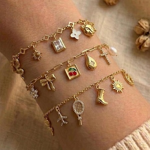 May include: Three gold-toned charm bracelets featuring various dangling pendants including a rectangular crystal, a red cherry motif, a sun, a cross, a bow, a cowboy boot, an airplane, a tennis racket, and a small flower shape with white stones.
