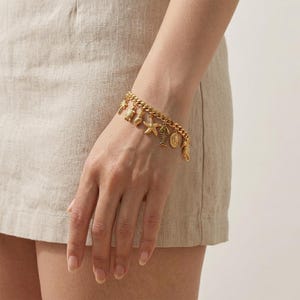 May include: A gold-toned chain link bracelet featuring multiple dangling charms, including a starfish, a fish, a seashell, and a circular medallion. The bracelet is constructed with a sturdy curb chain design and secures with a clasp, showcasing a polished metallic finish throughout the entire piece.