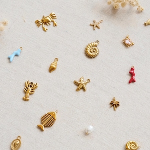 May include: A collection of small metal charms featuring various ocean-themed shapes including a crab, starfish, seashell, lobster, fish skeleton, and palm tree, alongside a light blue coral piece, a red coral piece, and a small white pearl bead.