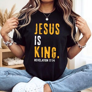 May include: A black short-sleeved t-shirt featuring the text JESUS IS KING in distressed yellow and white lettering, with the reference REVELATION 17:14 printed in smaller white text underneath the main graphic on the front of the garment.