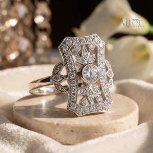 Puede incluir: Un anillo de tono plateado con una cara rectangular alargada y una piedra redonda central, rodeada de intrincada filigrana metálica y pequeñas piedras engastadas en pavé. El diseño incluye un borde milgrain y el texto Alter WhimsyCo.