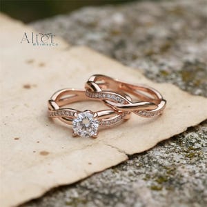 Könnte beinhalten: Zwei roségoldene Ringe, einer mit einem großen, klaren Mittelstein und der andere mit einem ähnlichen Design, werden auf einem Stück Papier gezeigt. Die Ringe haben ein verschlungenes Design mit kleinen, funkelnden Steinen. Der Markenname "Alter Whimsy Co" ist sichtbar.