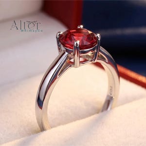 May include: A silver-toned ring featuring a round, faceted, vibrant red gemstone held in a four-prong setting. The band is smooth and polished. The text 'Alter WhimsyCo' is visible in the upper left corner of the frame.