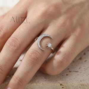 May include: A silver-toned open ring featuring a crescent moon shape adorned with small clear stones and a single star-shaped charm with a central clear stone. The ring has a polished metallic finish and a delicate band design.