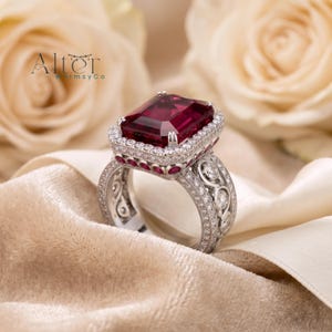 Peut inclure: Une bague argentée ornée d'une grande pierre précieuse rectangulaire rouge foncé au centre, entourée d'un halo de petites pierres blanches. L'anneau présente des détails métalliques complexes, des pierres blanches et des accents rouges.