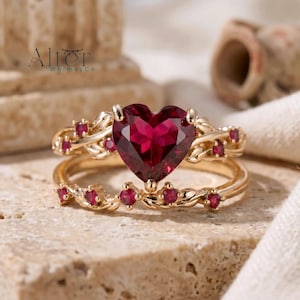 Puede incluir: Un juego de anillos de oro con una gema roja en forma de corazón. El anillo está adornado con gemas rojas más pequeñas y un delicado diseño en forma de vid. La marca "Alter Whimsy Co" es visible en la esquina superior izquierda.