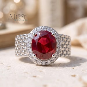 Puede incluir: Un anillo de tono plateado con una gran piedra preciosa de color rojo intenso y corte ovalado en el centro. La piedra central está rodeada por un halo de pequeñas piedras claras de corte redondo. La banda ancha tiene un patrón de metal tejido.