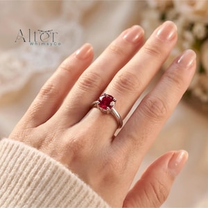 May include: A silver-toned ring featuring a round, faceted, deep red gemstone set in a four-prong setting. The ring is worn on a finger, and the text Alter WhimsyCo is visible in the upper left corner of the frame.