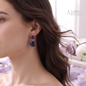 May include: A drop earring featuring two square-cut, deep purple amethyst gemstones set in silver-toned metal. The smaller stone sits at the earlobe, while the larger stone hangs below. The text Alter WhimsyCo is visible in the upper right corner.