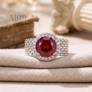 May include: A silver ring with a large, round, deep red gemstone at the center, surrounded by smaller white stones. The band has a woven texture. The ring is displayed on a beige fabric.
