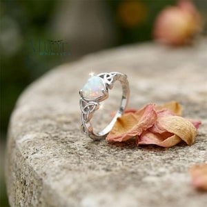 Puede incluir: Un anillo de plata con una piedra preciosa de ópalo en forma de pera. El anillo presenta diseños de nudos celtas en la banda y alrededor de la piedra preciosa. El ópalo tiene un aspecto blanco lechoso. El anillo se exhibe sobre una superficie de piedra con pétalos de flores secas.