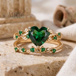May include: A gold ring set featuring a heart-shaped emerald center stone. The ring is accented with smaller emeralds and a delicate vine-like design. The set is displayed on a textured surface.