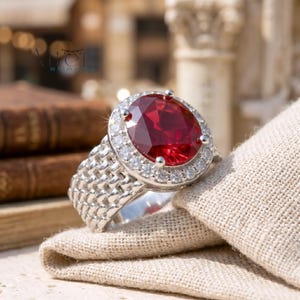 May include: A silver ring with a large, oval-cut, deep red gemstone surrounded by a halo of smaller, clear stones. The ring band has a textured, woven design. The ring is displayed on a beige cloth.