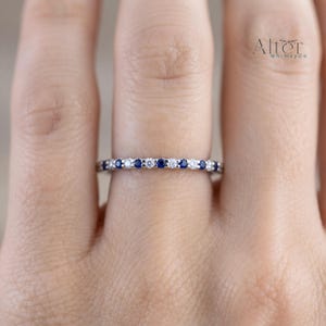 May include: A thin silver-toned band ring featuring an alternating pattern of small, round, deep blue sapphires and clear, sparkling white diamonds set in a straight line across the front. The text Alter WhimsyCo is visible in the upper right corner.