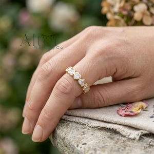 Puede incluir: Un anillo de tono dorado que presenta una banda continua de piedras transparentes en forma de corazón engastadas. El anillo se lleva en un dedo, mostrando el patrón repetitivo de corazones que rodea la banda. El metal tiene un acabado pulido.