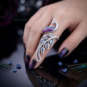 May include: A silver peacock ring with a colorful enamel design. The ring features a peacock with purple, blue, and silver details. The ring is on a finger with dark purple and black nail polish. The Alter Whimsy Co. logo is in the upper left corner.