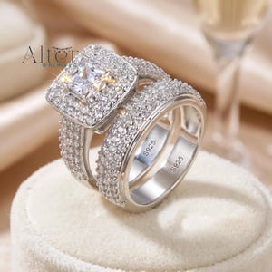 May include: A silver engagement ring set with a square-cut central stone and numerous smaller, sparkling stones. The band is also covered in smaller stones. The rings are displayed on a white surface. The inscription "S925" is visible on the inside of the bands.