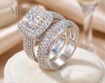 May include: A silver engagement ring set featuring a square-cut center stone surrounded by smaller, sparkling stones. The band is also encrusted with smaller stones. The rings are displayed on a white surface. The inscription "S925" is visible on the inside of the bands.