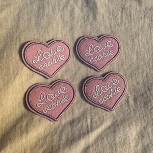May include: Four pink heart-shaped patches with embroidered text that reads "Love cachie" in white cursive. Each patch has a purple border. The patches are arranged on a light beige fabric.