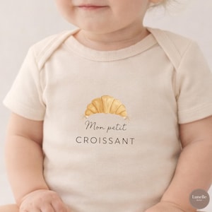 May include: A cream-coloured baby bodysuit featuring a watercolour illustration of a croissant and the text "Mon petit CROISSANT" in black. The design is centred on the front of the short-sleeved onesie.
