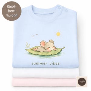 May include: A stack of three baby shirts: light blue, white, and pink. The top shirt features a cartoon mouse on a leaf with the text "summer vibes". The mouse is tan with pink ears, and the sun is yellow. The shirts are soft and folded.
