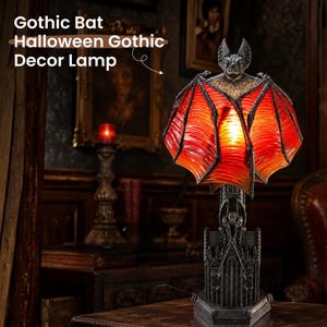 May include: A table lamp featuring a bat with outstretched wings that serve as a red and orange translucent shade. The base is sculpted to resemble a dark gray gothic cathedral with architectural details and a small gargoyle figure at the center.