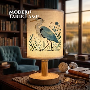May include: A modern table lamp with a cylindrical lampshade featuring a blue heron illustration amidst floral elements. The lamp has a wooden base and stem. The text "MODERN TABLE LAMP" is displayed on the upper left.