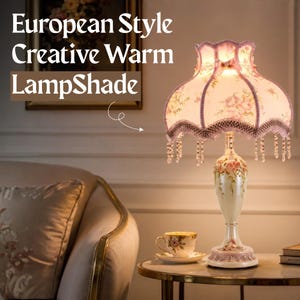 May include: A table lamp with a cream-colored ceramic base featuring pink floral accents and a scalloped fabric lampshade with pink roses, purple trim, and hanging crystal beads. The text European Style Creative Warm LampShade is visible.