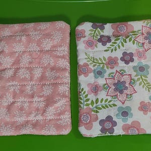 May include: Two rectangular fabric items. One is pink with white floral patterns and horizontal stitching. The other is white with pink, purple, and blue floral patterns and green leaves. Both are on a green background.