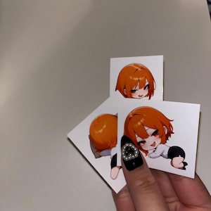 以下が含まれることがあります： オレンジ色の髪と赤い目の漫画のキャラクターが描かれた、3つの白い正方形のステッカー。キャラクターは、白黒の衣装で、さまざまなポーズで描かれています。ステッカーは、宝石が付いた黒い爪の手で持たれています。