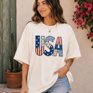 May include: Cream-colored oversized t-shirt with the word "USA" in a patriotic design. The letters feature red and white stripes, blue gingham, and stars. The shirt is paired with denim shorts.