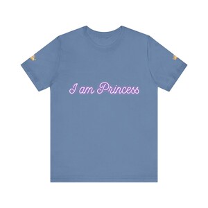 May include: A light blue t-shirt with the text "I am Princess" in a cursive font. The shirt has short sleeves and a crew neck. A small gold crown is printed on the left sleeve.