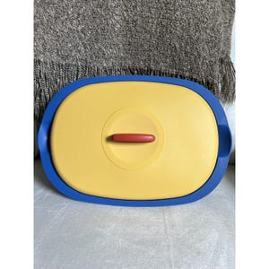 May include: A yellow and blue plastic container with a red handle. The container has a blue rim and a yellow lid with a red handle. The container is on a white surface.