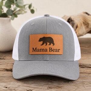 May include: A grey trucker cap with a white mesh back and a brown leather patch. The patch features a black bear silhouette and the words "Mama Bear". The cap is on a wooden surface.