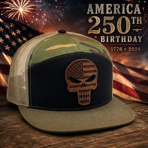 Puede incluir: Gorra de camionero camuflaje y negra con visera marrón. La gorra presenta un parche de cuero con un diseño de calavera que incorpora la bandera estadounidense. El texto "AMERICA 250TH BIRTHDAY 1776 * 2026" es visible en el fondo.