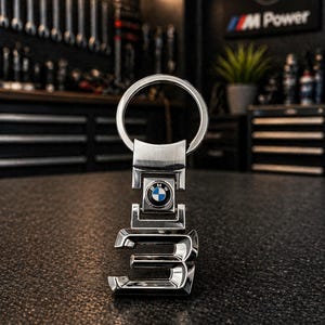 May include: A shiny silver metal keychain featuring the number "3" and the BMW logo. The keychain has a circular ring for keys and is set against a dark, textured surface. The background includes tools and a BMW sign.