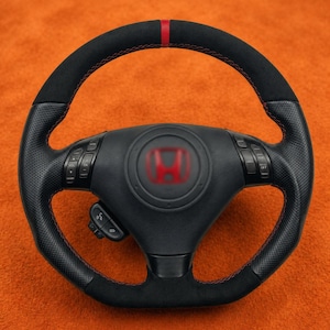 May include: Black steering wheel with red stitching and a red stripe at the top. The center features a red Honda logo. The wheel has perforated leather sections and control buttons.