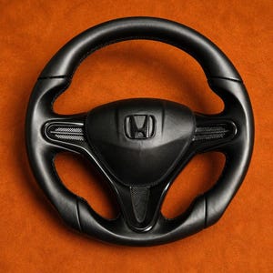 May include: A black steering wheel featuring the Honda logo at its centre. The wheel has a leather-like texture with visible stitching. The background is a solid orange colour.