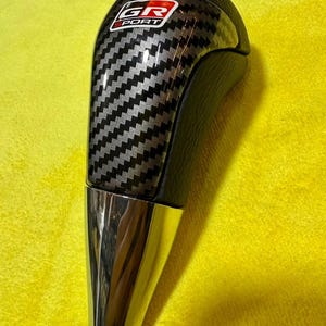 Puede incluir: Un pomo de cambio negro y plateado con un patr&oacute;n de fibra de carbono. La parte superior del pomo tiene el texto "GR SPORT" en un logotipo rojo y blanco. La base es cromada.