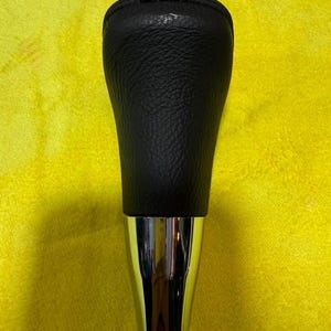 Puede incluir: Un pomo de palanca de cambios de cuero negro con base cromada. La parte superior es negra y texturizada, mientras que la base es de color plateado brillante. El pomo de la palanca de cambios est&aacute; sobre un fondo amarillo.