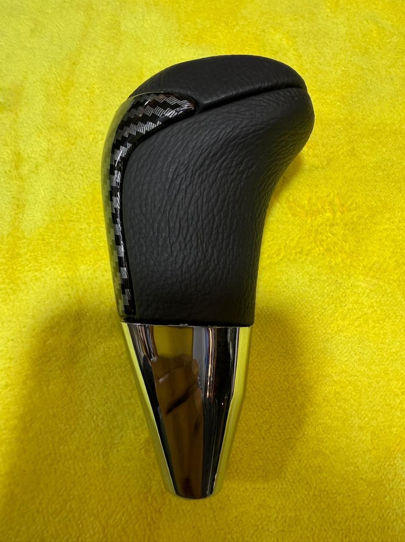 Puede incluir: Pomo de palanca de cambios de coche negro y plateado con una parte superior de cuero texturizado y un acento de fibra de carbono. La base es cromada. La palanca de cambios est&aacute; sobre un fondo amarillo.