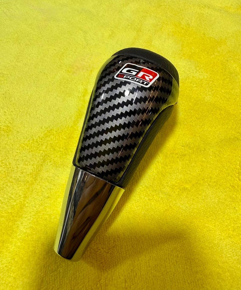 Puede incluir: Pomo de palanca de cambios con un patr&oacute;n de fibra de carbono negro y plateado y una base cromada. La parte superior del pomo presenta el logotipo "GR SPORT" rojo y blanco.