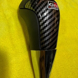 Puede incluir: Pomo de cambio de fibra de carbono negra y plateada con base cromada. La parte superior del pomo es de cuero negro. El pomo de cambio tiene el texto "GR SPORT" en un logotipo rojo y blanco.