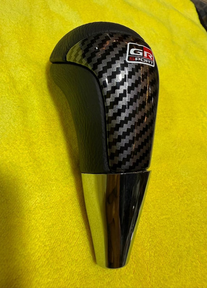 Puede incluir: Pomo de cambio de fibra de carbono negra y plateada con base cromada. La parte superior del pomo es de cuero negro. El pomo de cambio tiene el texto "GR SPORT" en un logotipo rojo y blanco.