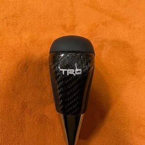 May include: A black and silver TRD gear shift knob. The top is black, the middle section features a carbon fibre weave pattern with the TRD logo, and the bottom is silver. Designed for a vehicle's gear shift.