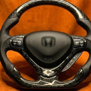 May include: A black and carbon fiber steering wheel with a Honda logo in the center. The wheel has perforated leather grips and control buttons. The carbon fiber has a unique, marbled pattern.